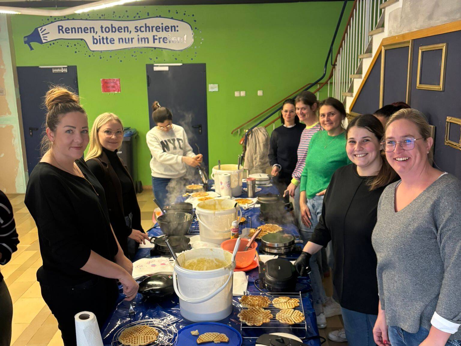 Neun Frauen auf dem Bild stehen um einen blauen Tisch und backen Waffeln