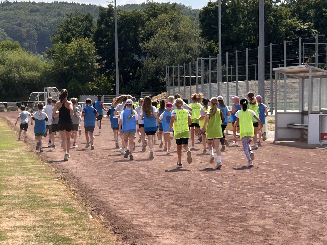 Hier sind Personen, vorwiegend Kinder von der Rückseite zu sehen, die auf einem Ascheplatz laufen, bei sonnigem Wetter.